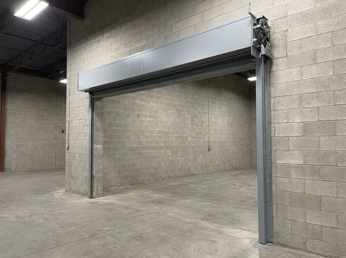A rolling steel fire door rolled up into its boxed steel hood above an opening in a concrete block fire wall, with side guides running down both jambs and the release governor and its chain on the end bracket